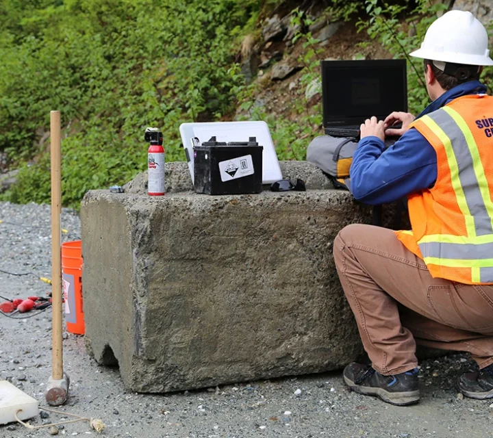 Surveyor in hard hat and vest works on a laptop outdoors, near equipment on a concrete block.