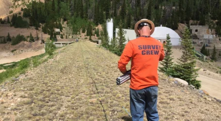 Survey crew member in orange shirt working outdoors near forested area with structures in the background.