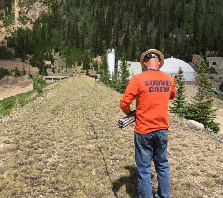 Survey crew member in orange shirt working outdoors near forested area with structures in the background.