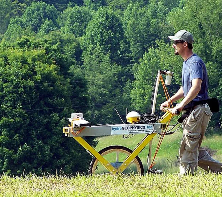 Technician operating wheeled electromagnetic induction system across grassy field to map subsurface conductivity variations and buried features for site characterization and planning using electromagnetic induction geophysical survey methods.
