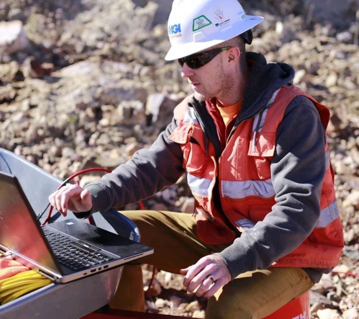 Geophysicist reviewing seismic refraction data on a field laptop during excavation rippability analysis, interpreting subsurface velocity results to evaluate rock strength, fracture presence, and excavation resistance across the site.