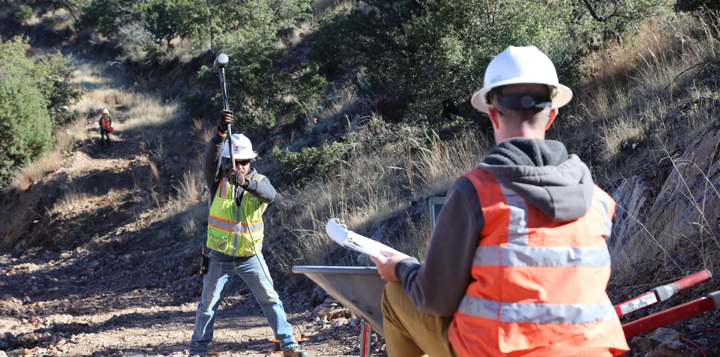 Two workers outdoors performing seismic testing; one swings a sledgehammer, the other monitors with equipment and notes.