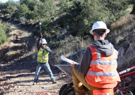 Two workers outdoors performing seismic testing; one swings a sledgehammer, the other monitors with equipment and notes.