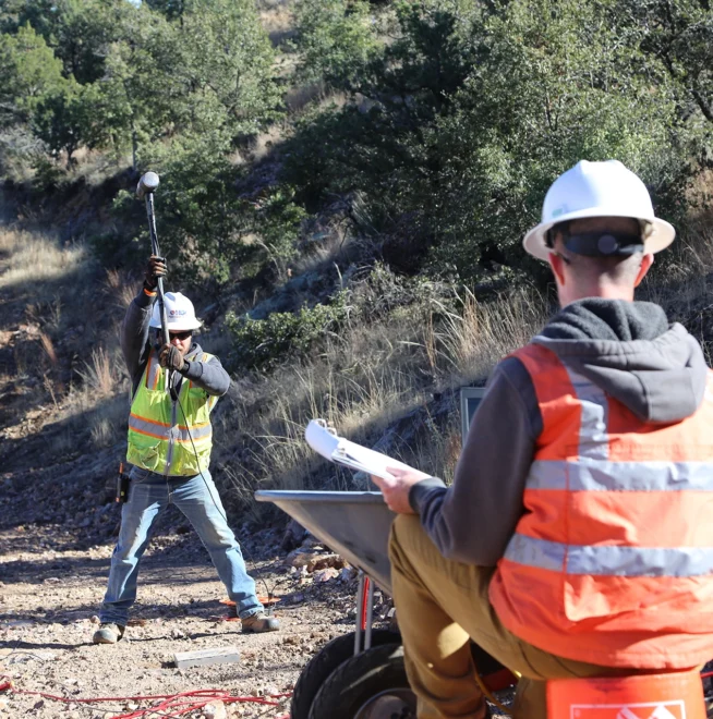 Two workers outdoors performing seismic testing; one swings a sledgehammer, the other monitors with equipment and notes.