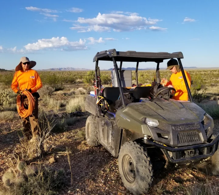 Geophysical field crew conducting geohazard characterization survey in desert terrain with off-road vehicle and surface instrumentation, collecting subsurface data to assess faults, unstable materials, and potential geological hazards before construction.