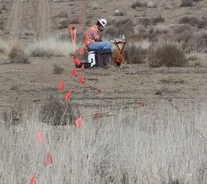Technician conducting geohazard characterization along flagged seismic line in desert terrain, seated near surface cables and sensors used to image subsurface hazards such as voids, fractured bedrock, and unstable ground conditions.