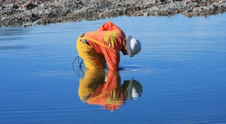Person in orange gear bends over water in rocky area, creating ripples.