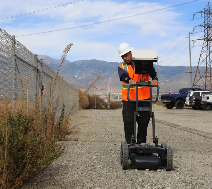 Worker in safety gear uses ground-penetrating radar near a fence with mountains in the background.