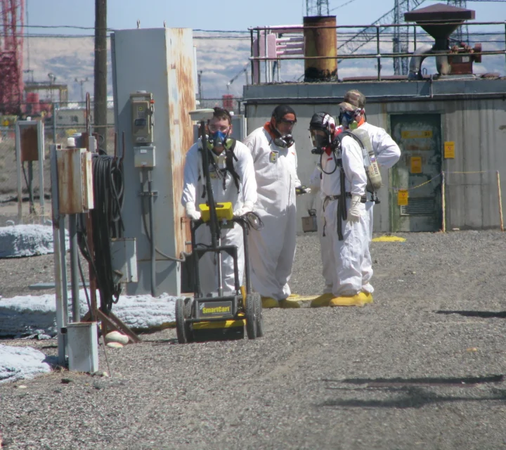 People in protective suits and masks using equipment outdoors at an industrial site.