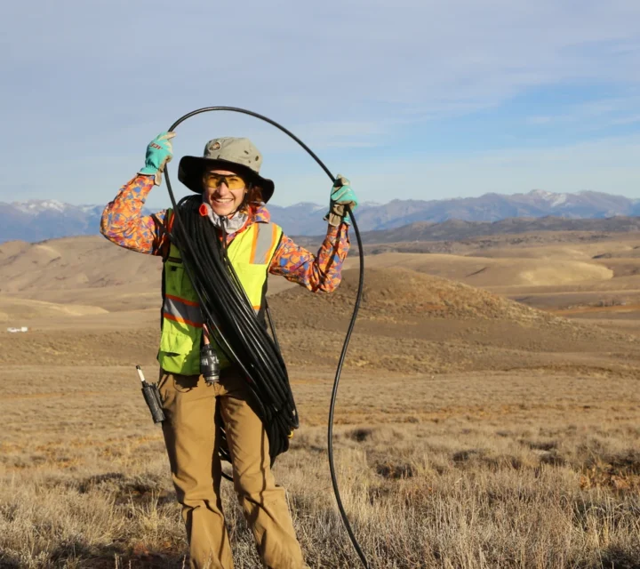 Person in high-vis gear holding a cable in a vast, grassy landscape with mountains in the background.