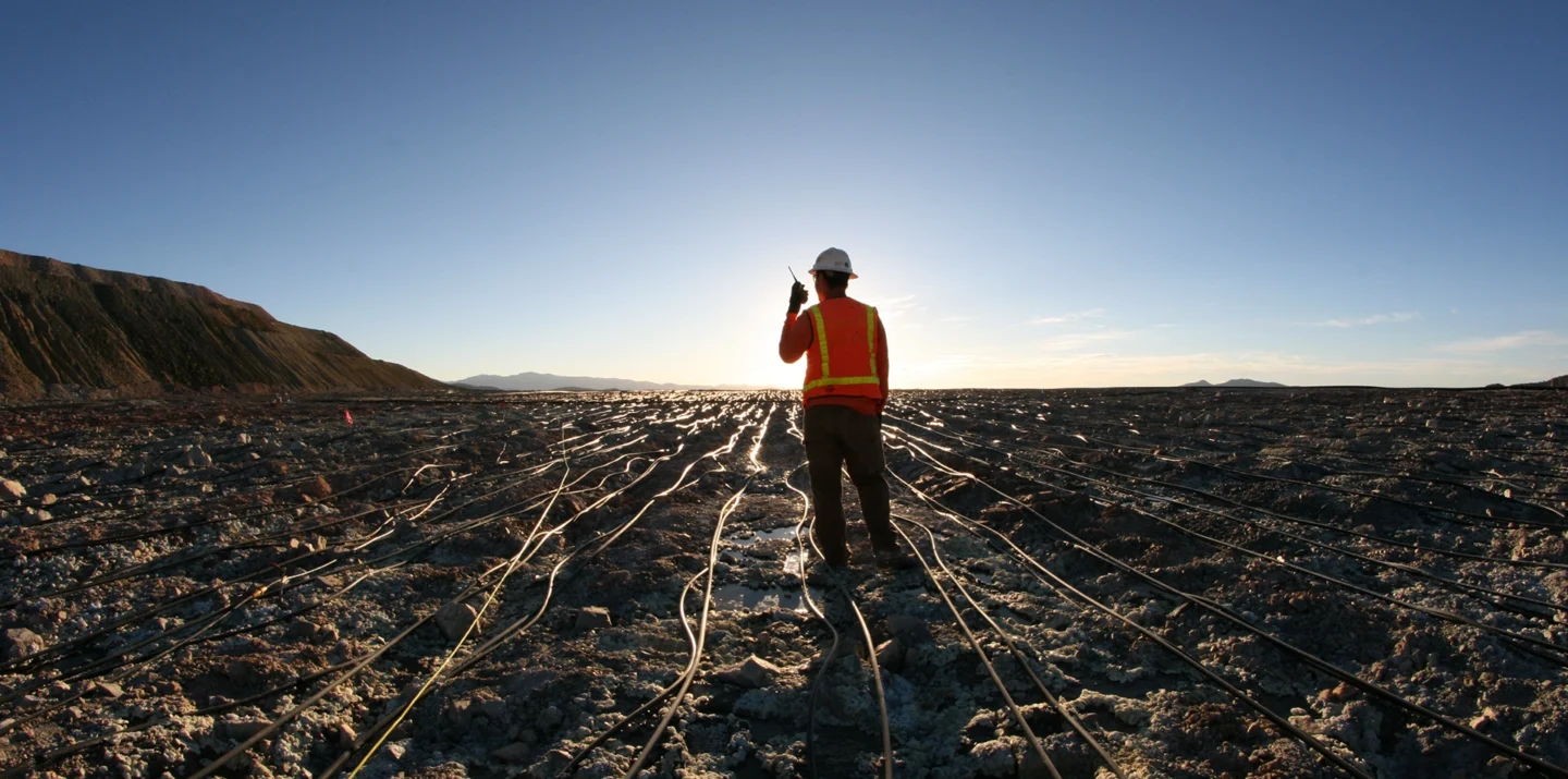 Worker in a hard hat and vest on a barren, wired landscape with a bright sky background.
