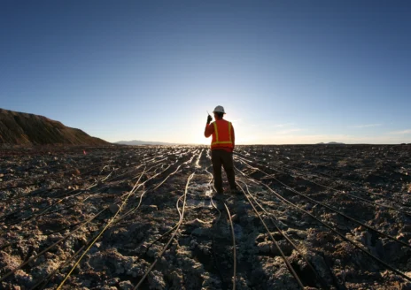 Worker in a hard hat and vest on a barren, wired landscape with a bright sky background.