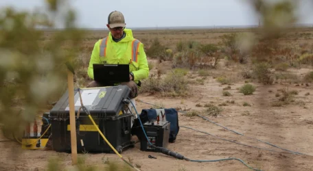 Person in high-visibility clothing sits outdoors with equipment, working on a laptop in a desert-like area.
