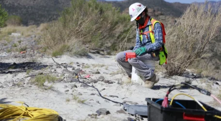 Worker in safety gear kneels on sandy ground handling cables with mountains in the background.