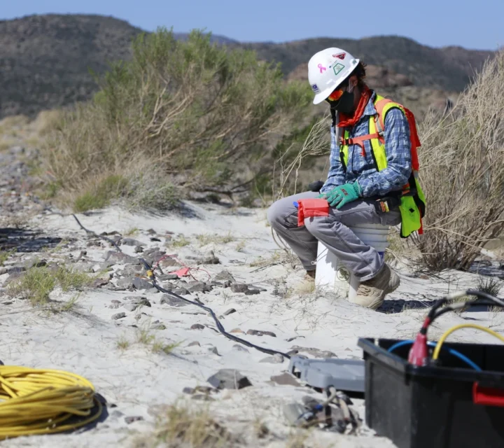 Worker in safety gear kneels on sandy ground handling cables with mountains in the background.