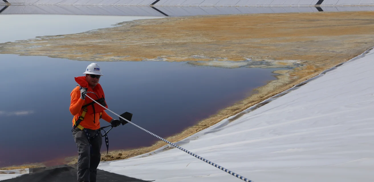 Worker in orange gear inspects large, partially filled industrial pond with safety harness and rope.