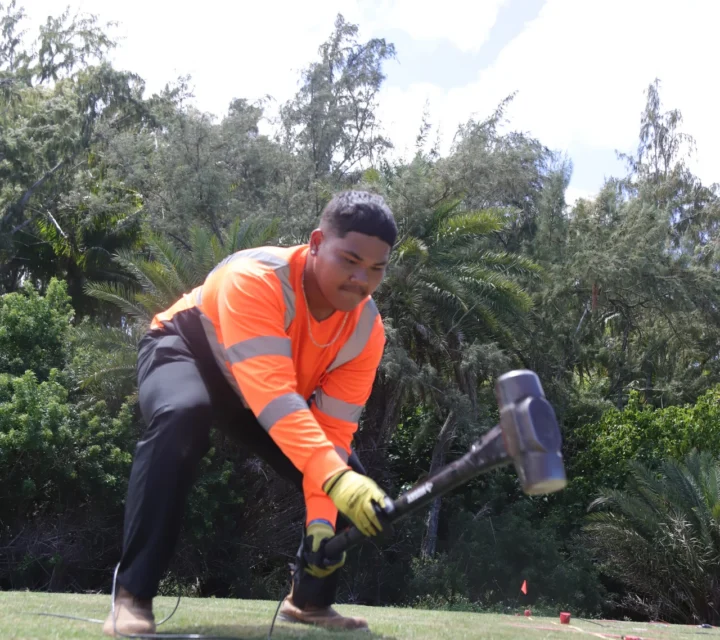 Technician striking ground with hammer source during MASW surveys while geophone array records surface waves to analyze dispersion and estimate shear-wave velocity variations with depth across near-surface profile.