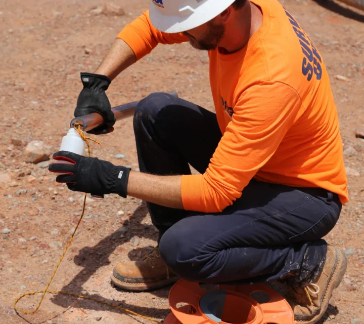 Construction worker kneeling with measuring tool on sandy ground wearing hard hat, gloves, and orange safety shirt.