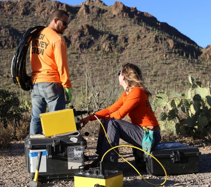 Two people in orange shirts with geology equipment in a desert landscape.