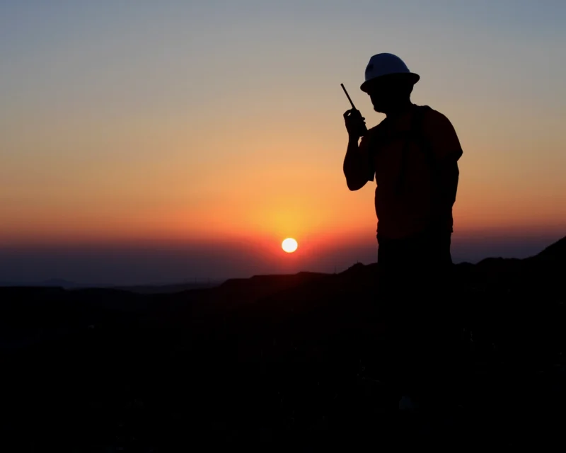Silhouette of a person in a hard hat using a walkie-talkie against a sunset backdrop.
