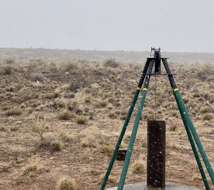 Tripod well casing in a barren, grassy landscape under a cloudy sky.