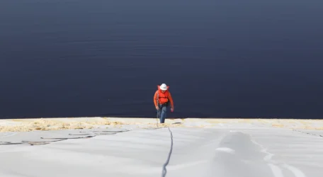 Person in a red jacket and hat climbing a steep white surface beside a dark water body.