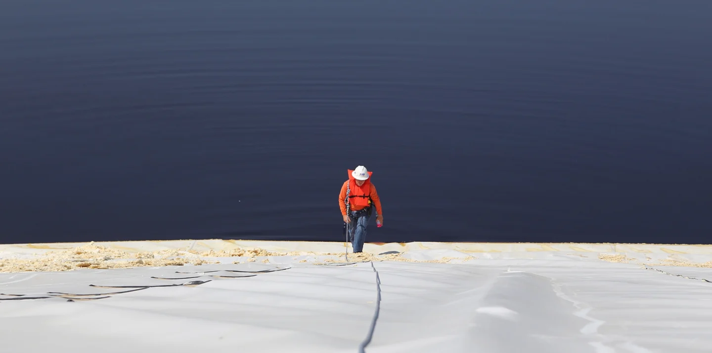 Person in a red jacket climbing a steep surface with a rope near a vast, calm body of water.