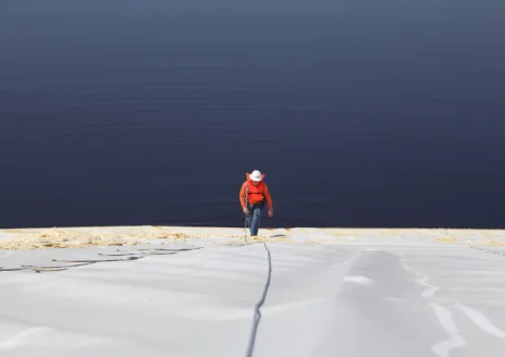 Person in a red jacket climbing a steep surface with a rope near a vast, calm body of water.