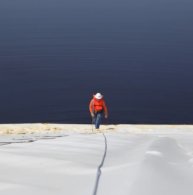 Person in a red jacket climbing a steep surface with a rope near a vast, calm body of water.