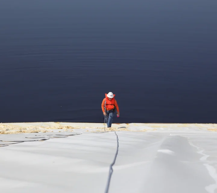 Technician walking along a geomembrane-lined pond during pond leak detection survey to evaluate liner seams and identify potential breach locations.