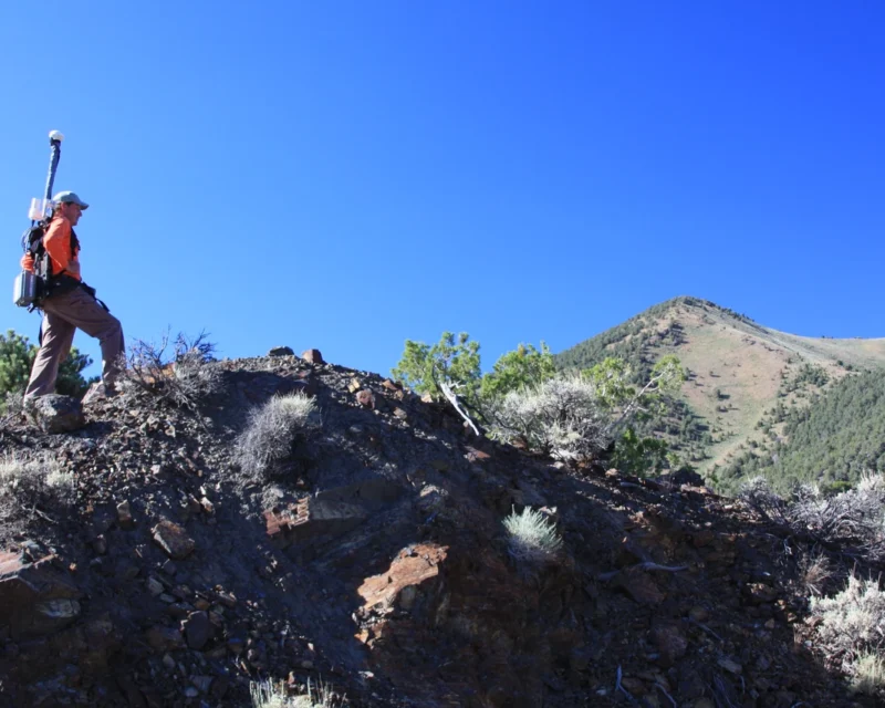 Field technician carrying magnetometer across rocky hillside during potential field methods survey to measure natural magnetic variations and map subsurface structure and lithologic contrasts across regional terrain.