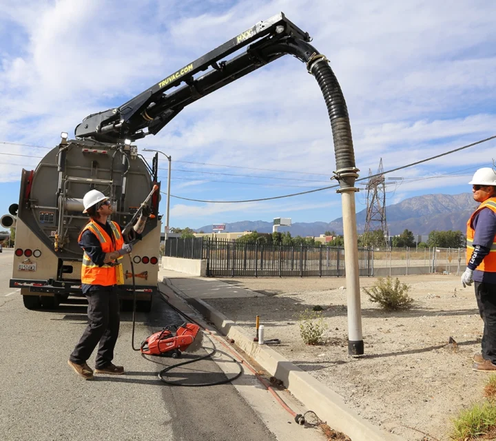 Workers in safety gear operate a vacuum truck to clean drainage on a roadside.