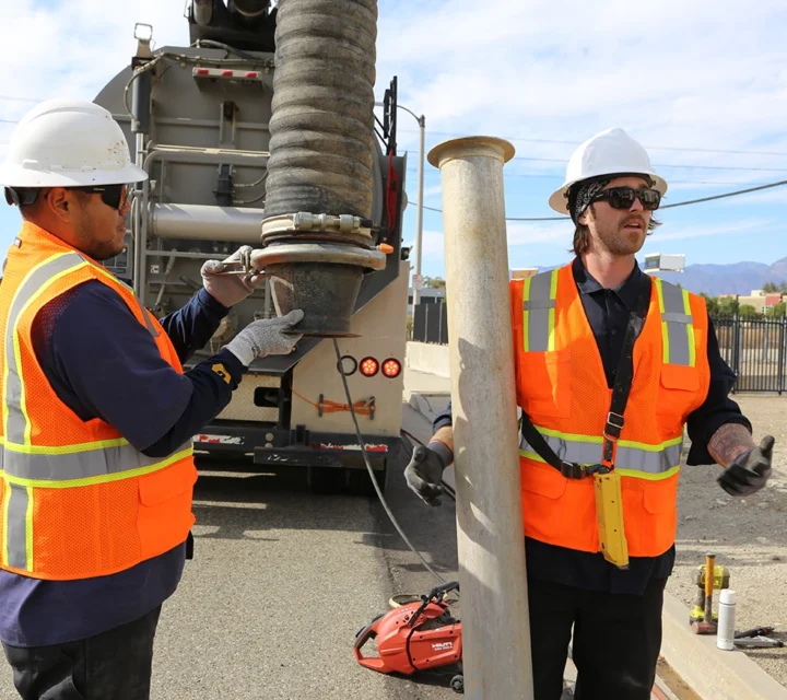 Two workers in safety gear operate vacuum equipment on a street.