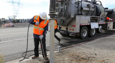 Worker in safety gear uses equipment near utility truck on roadside.