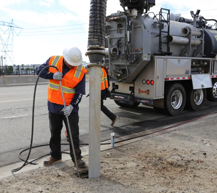 Worker in safety gear uses equipment near utility truck on roadside.