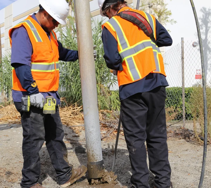 Two construction workers in safety gear operate equipment for a utility pole installation.