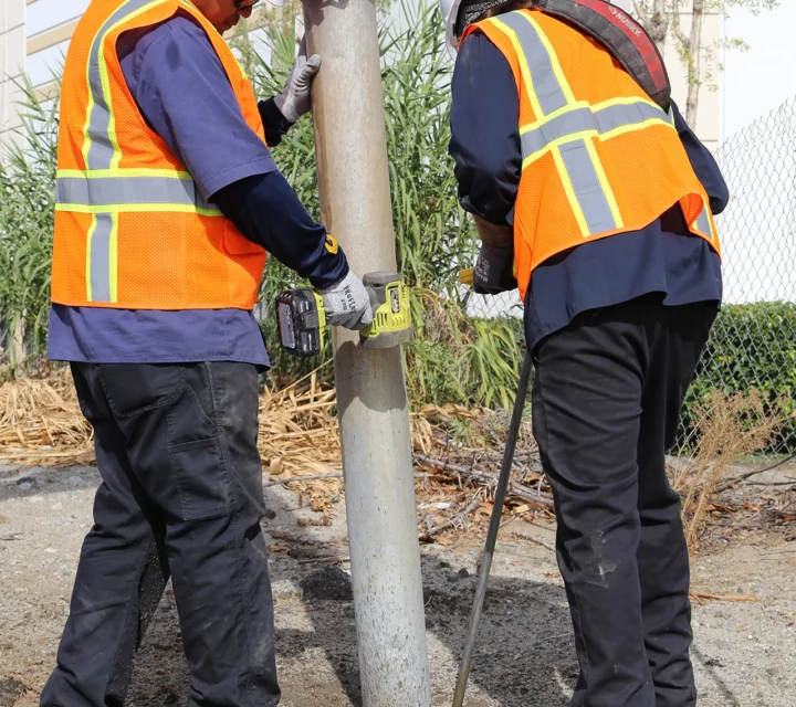 Two workers in safety gear securing a pole outside a building with a drill and crowbar.