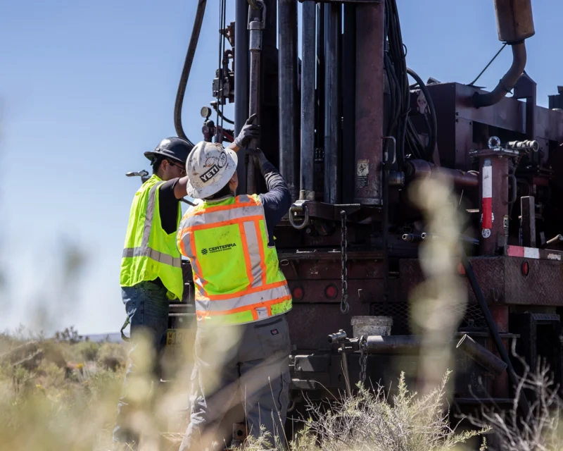Two workers in safety gear operate heavy drilling equipment outdoors.