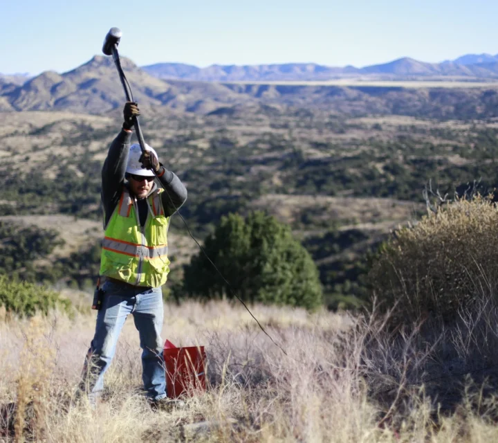 Technician using hammer source and geophone cable during seismic methods survey to measure wave travel through subsurface materials for characterization of structure, stiffness, and depth variations across rugged terrain.