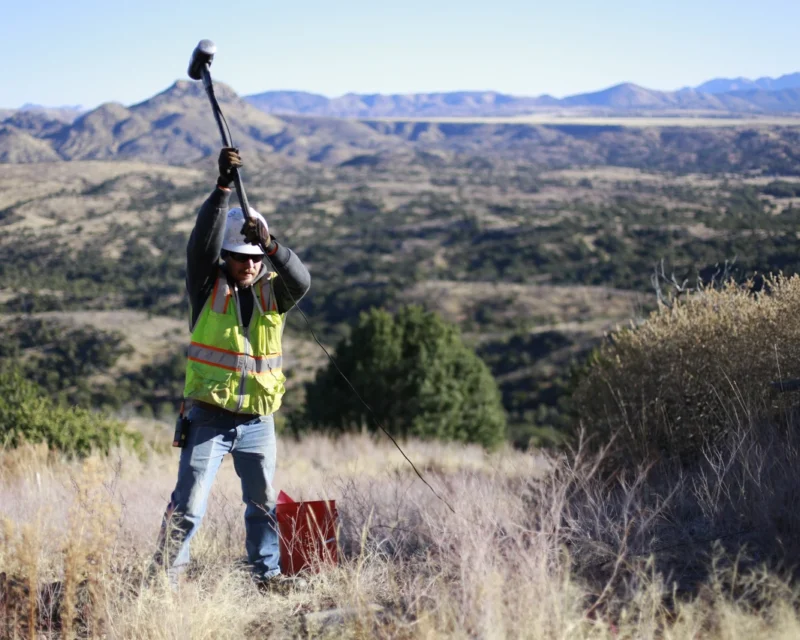 Technician using hammer source and geophone cable during seismic methods survey to measure wave travel through subsurface materials for characterization of structure, stiffness, and depth variations across rugged terrain.