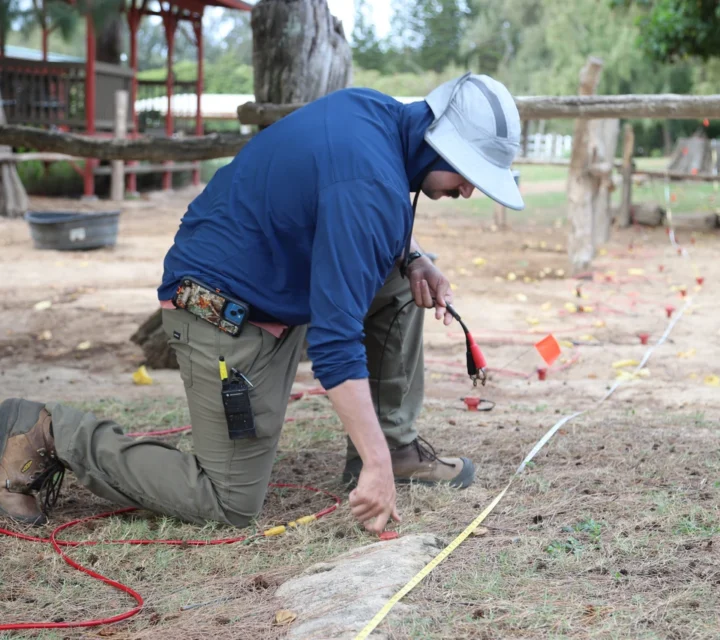 Person kneeling outdoors measuring ground with a tape, surrounded by flags and cables.
