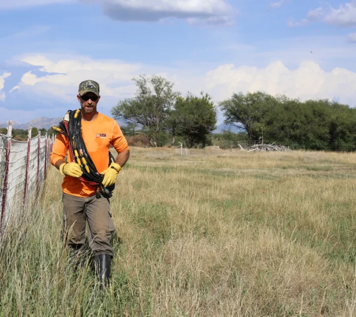 Man in orange shirt and boots carrying equipment, walking beside a wire fence in a grassy field under a blue sky.