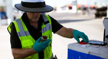 Worker in safety gear examining a sample jar beside a cooler outdoors.