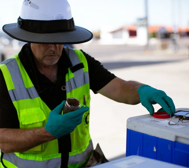 Worker in safety gear examining a sample jar beside a cooler outdoors.