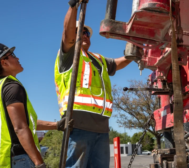 Construction workers in safety gear operate heavy machinery outdoors, wearing bright yellow vests and hard hats.