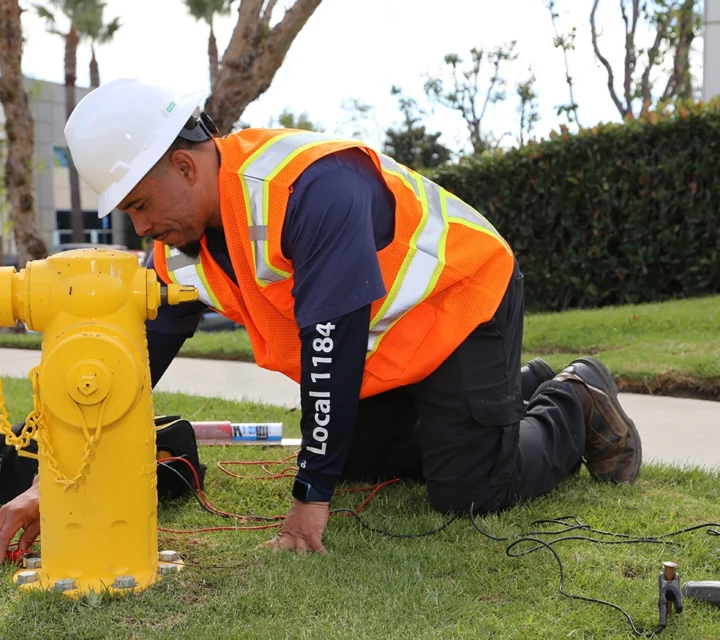 Worker in safety gear and helmet examines a yellow fire hydrant on a grassy area.