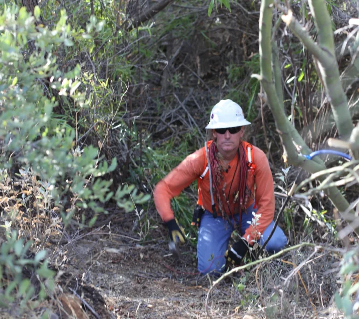 Geophysical technician kneeling in vegetated drainage area conducting subsurface profiling survey to map contamination pathways, moisture variations, and subsurface material contrasts using non-invasive surface geophysical methods.