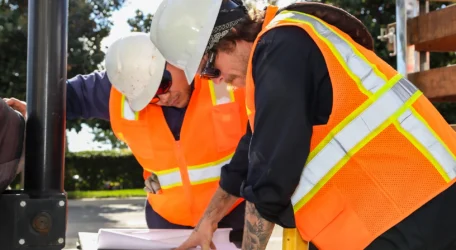 Two construction workers in orange vests and helmets study blueprints outdoors.