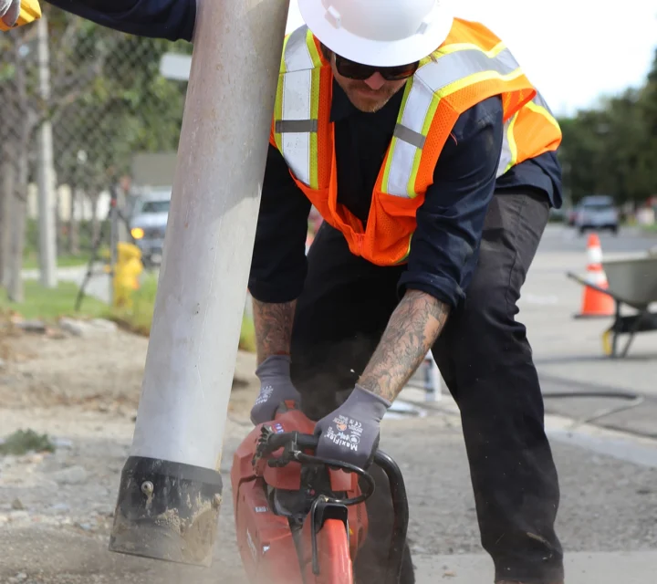 Worker in safety gear cutting road surface with a saw near a pole.