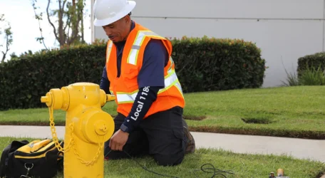 Worker in safety gear inspecting a yellow fire hydrant on a grassy area.